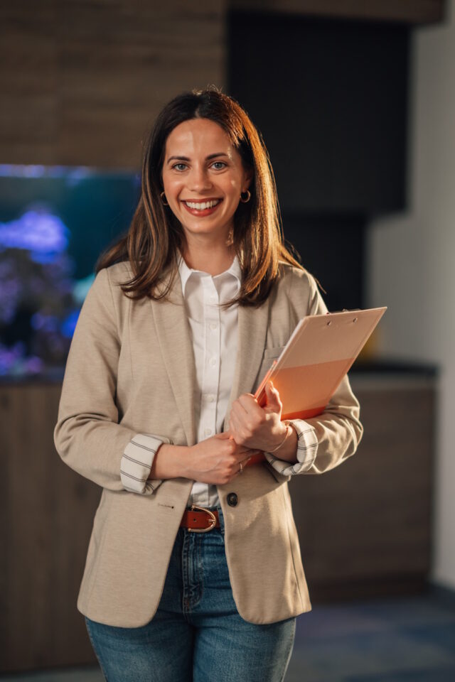 Corporate company worker in business casual outfit standing at modern office with clipboard in hands and smiling at camera. Smart casual dressed businesswoman with clipboard posing at enterprise.